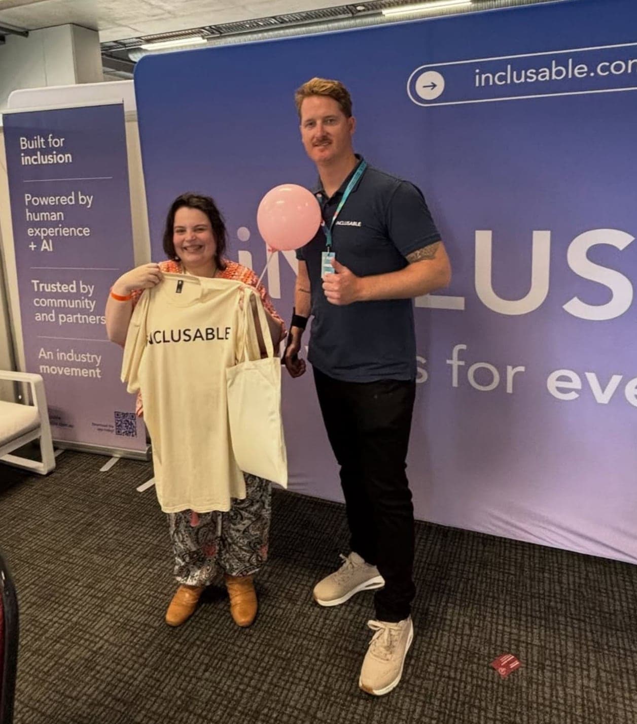 Founder Brodie Duncan and a community member smiling at the Inclusable expo stand, holding an Inclusable t-shirt and a pink balloon in front of the brand wall
