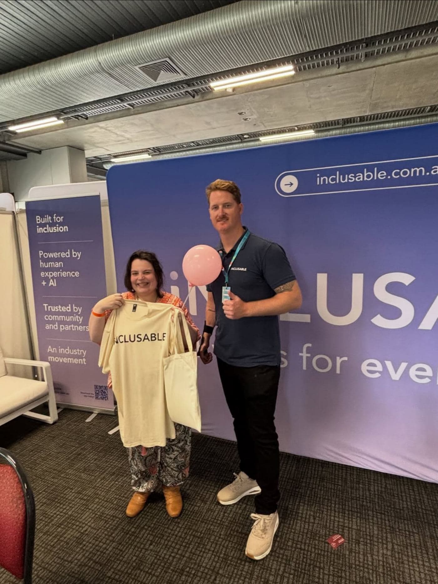 Founder Brodie Duncan and a community member smiling at the Inclusable expo stand, holding an Inclusable t-shirt and a pink balloon in front of the brand wall
