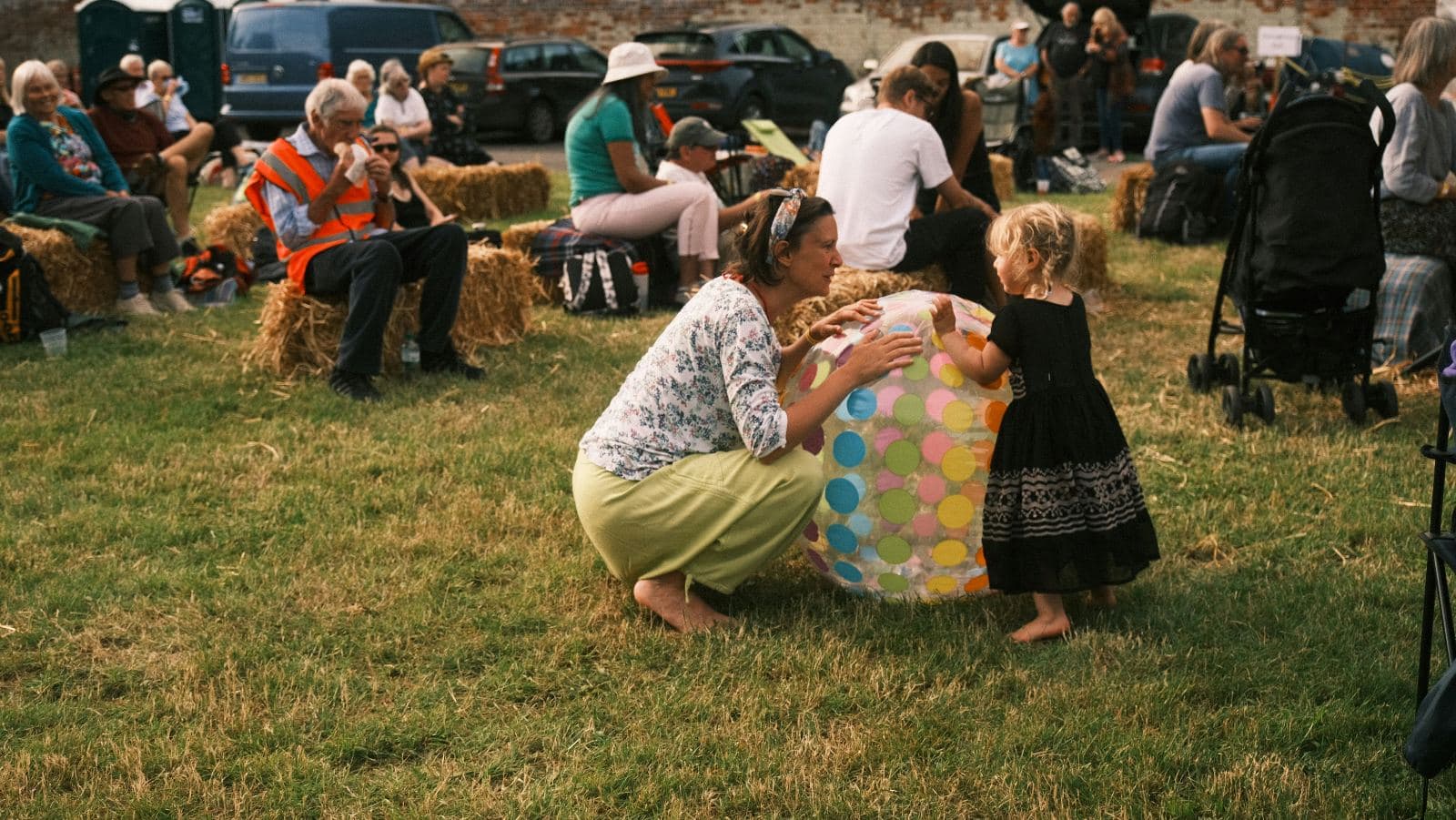 A relaxed community festival on grass with hay-bale seating — a mother plays with a toddler and a polka-dot ball while people of all ages watch on