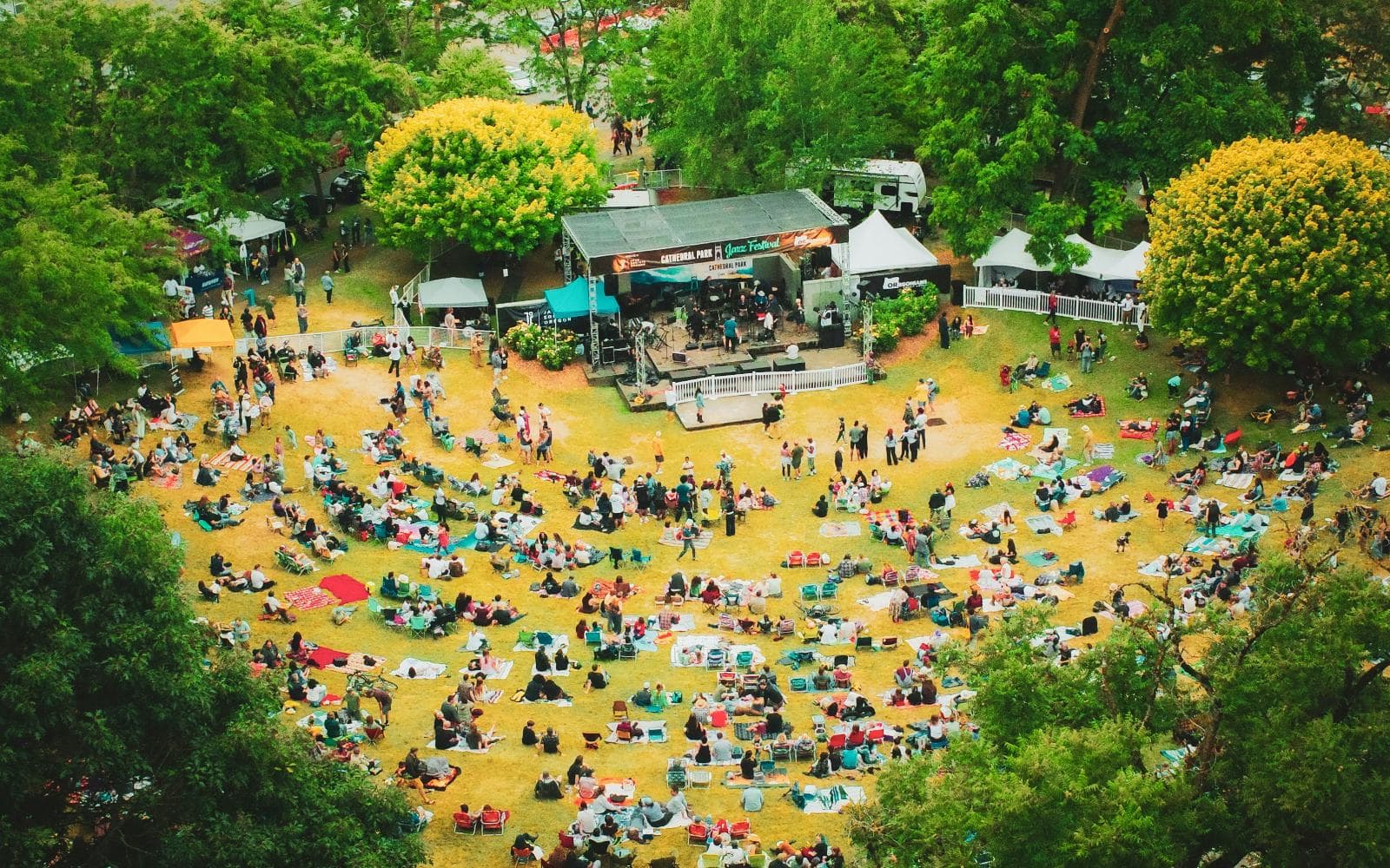 An aerial view of an outdoor festival in a leafy park, with hundreds of people on picnic rugs and chairs around a stage
