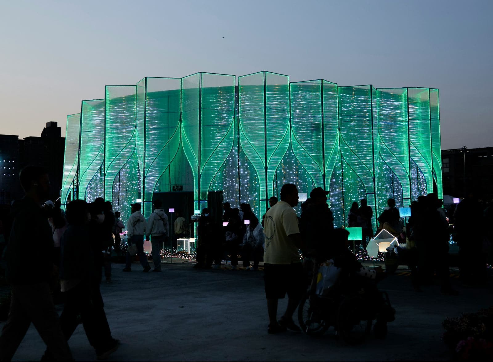 People at a glowing light-festival pavilion at dusk, including a person pushing a wheelchair in the foreground