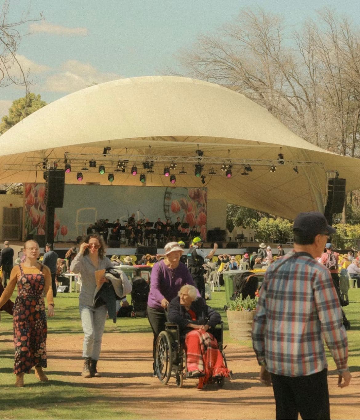A woman pushing an older woman in a wheelchair through a sunny outdoor festival, with a covered stage behind them