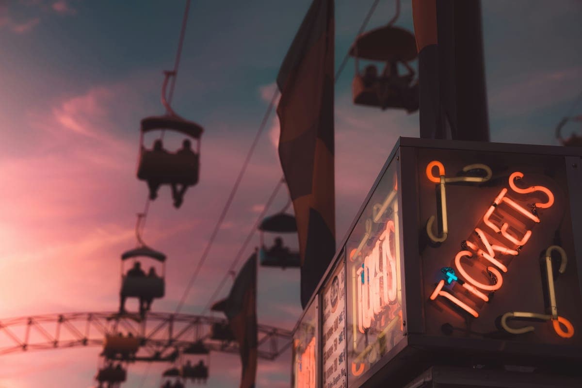 A glowing neon ‘Tickets’ sign at a fairground at dusk, with a chairlift and rides against a pink sunset sky