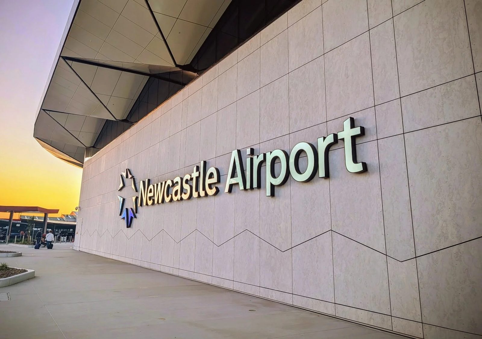 The Newcastle Airport sign and logo on the terminal’s outside wall at sunset, with travellers wheeling bags along the wide, level footpath