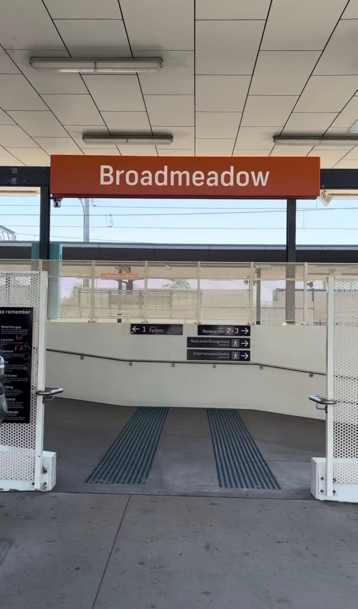 Entrance to Broadmeadow Station under the station sign: a ramp with tactile strips leads down, with signs to Platform 1 for Sydney and Platforms 2 and 3 for Newcastle